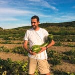 Homme tenant une grande courge dans un potager, sous un ciel bleu en fin de journée.