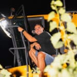 Homme souriant assis sur une machine de récolte, de nuit, entouré de vignes.