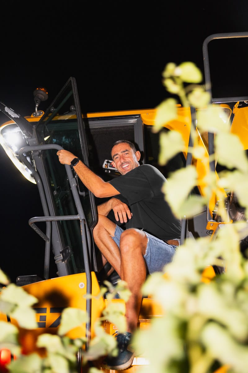 Homme souriant assis sur une machine de récolte, de nuit, entouré de vignes.