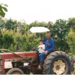 Homme et enfant assis sur un tracteur rouge dans un verger, entourés d'arbres.