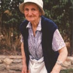 Femme âgée souriante portant un chapeau et s'appuyant sur une canne, en extérieur.