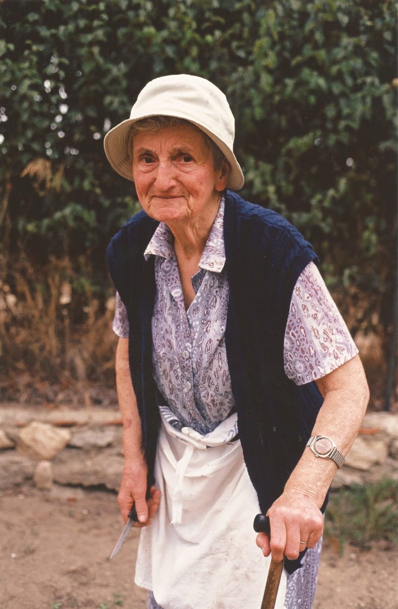 Femme âgée souriante portant un chapeau et s'appuyant sur une canne, en extérieur.